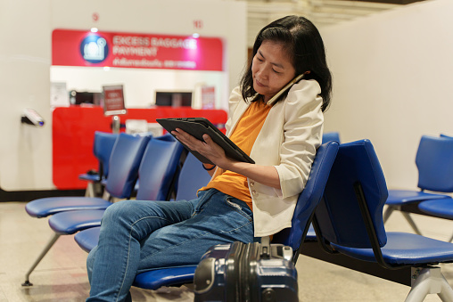 Young Asian business woman using laptop computer checking business trip plan and flight time schedule confirming meeting document with colleague before departure when waiting for flight departure at international airport terminal gate.
