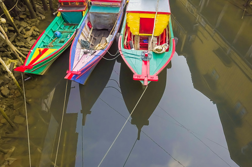 Many small passenger ships is at anchor between wooden buildings in shallow water. Colorful fishing boats in harbor during the day. Small engine boats.