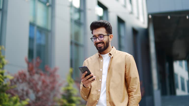 A young smiling businessman is using a smartphone while walking along the street near an office building
