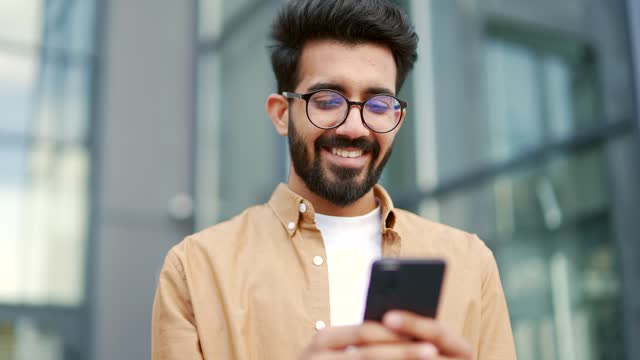 Close up. A young smiling man is using a smartphone while standing on the street near an office building