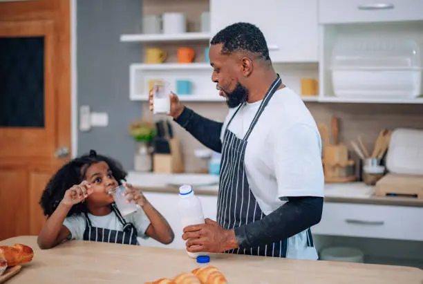 In the kitchen, a joyful father and his playful daughter bond, laugh, and cheer during a milk-drinking competition. They develop a precious memory, enjoying quality time and supporting each other.