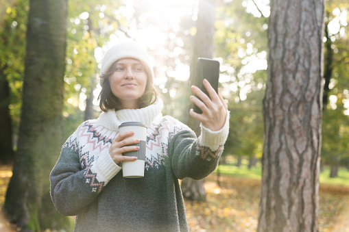 A woman is holding a phone and a cup of hot drink. Woman taking selfie on smartphone