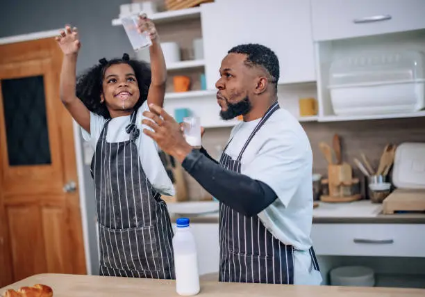 In the kitchen, a joyful father and his playful daughter bond, laugh, and cheer during a milk-drinking competition. They develop a precious memory, enjoying quality time and supporting each other.