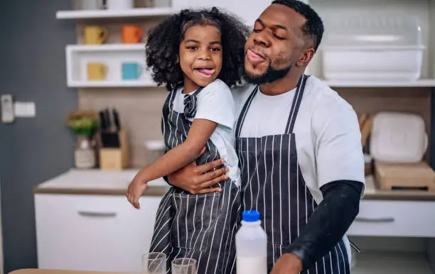 In the kitchen, a joyful father and his playful daughter bond, laugh, and cheer during a milk-drinking competition. They develop a precious memory, enjoying quality time and supporting each other.