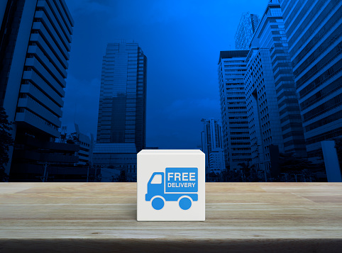 Free delivery truck icon on white block cube on wooden table over modern office city tower and skyscraper, Business transportation service concept