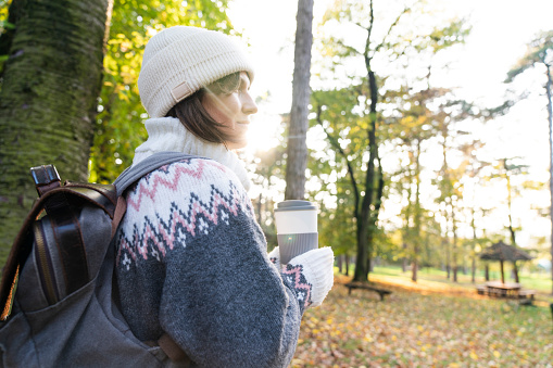 Woman traveler with backpack is holding a cup of hot drink in autumn park.