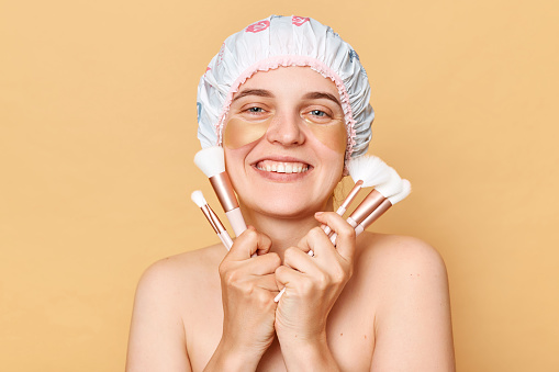 Joyful woman wearing shower cap standing isolated over beige background posing with big variety of makeup brushes doing beauty routine procedures after taking shower.