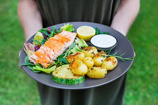 Woman hands holding baked salmon fillet with potatoes and vegetables in the garden summer time.