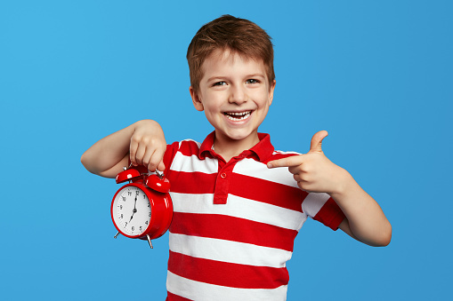 Cheerful cute child in casual red striped shirt, pointing index finger on red alarm clock. Posing isolated on blue background studio portrait. People emotions lifestyle concept. Mock up copy space.