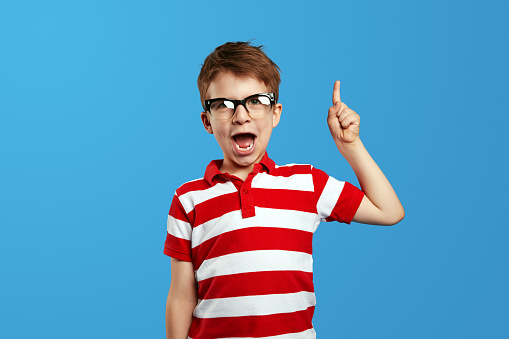 Little smart nerdy boy in eyeglasses and red striped shirt shouting and pointing up against blue background. Studio portrait of cute preschool child