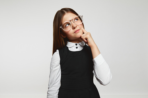 Thoughtful girl in glasses and school uniform touching cheek and looking up while thinking against white background.