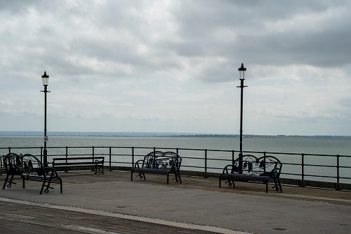 Whitby, UK. June 19, 2021.  A couple sit on a bench with an ornate lamppost to one side.  A pier with a lighthouse is in the middle distance and a sky with cloud is above.