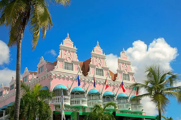 The pink Dutch style Royal Plaza with two palm trees at the side. It is set off with white clouds and blue sky. This building is an icon for Oranjestad.