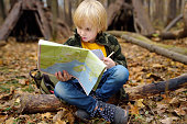 Little boy scout is orienteering in forest. Child is sitting on fallen tree and looking on map on background of teepee hut.