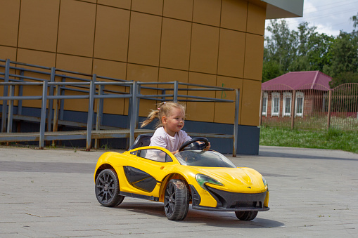 Beautiful baby girl smiling while sitting in an electric yellow car in the summer in the park. The wind blows the hair of a child riding an electric car in summer. Happy childhood concept..