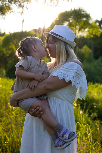 In the summer, against the backdrop of a sunset in nature, a mother gently holds her baby daughter in her arms. Mom and daughter are together and touch each other's noses. Mom and baby concept..