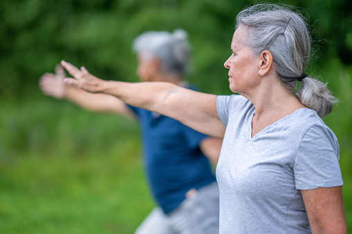 A senior couple take their Tai Chi outdoors on a warm summers day as they enjoy the fresh air.  They are both dressed casually and are focused on their movements and breathing.
