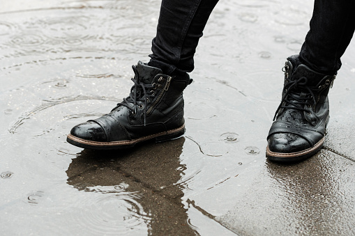 Black leather boots on a sidewalk puddle. Unrecognizable person wearing black boots and trousers. Bad weather concept.