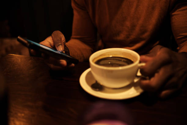 Unrecognizable black male using phone and drinking coffee sitting on a table. We can only see his hands, it is a dark moody image in a cozy pub.