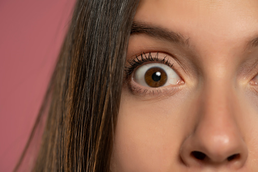 The wide open brown eye of a young woman: a concept of surprise and close observation on a pink background