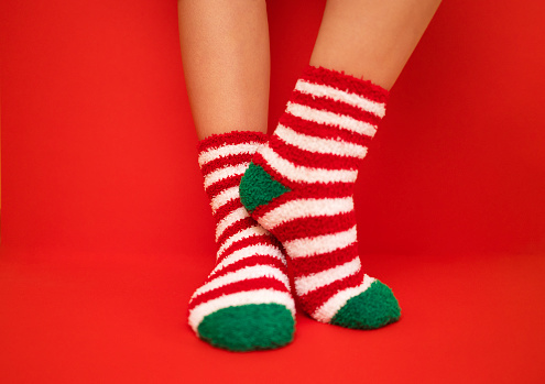Female feet in fluffy New Year or Christmas warm socks. The colors of the socks are red and white stripes and green heels and tips. Girls legs on a red bright background. Celebration holiday concept.