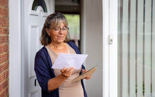Mature woman at the front door of her house checking her mail and reading an envelope - domestic life concepts