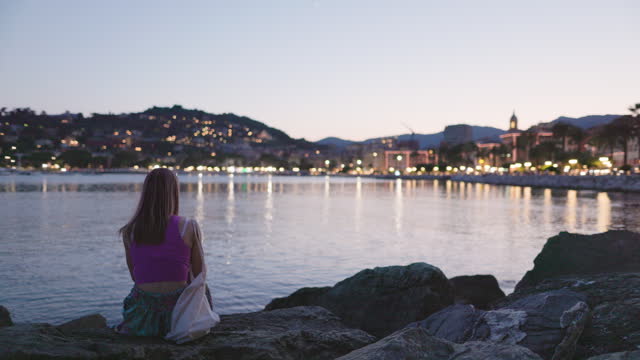Teenager girl sitting on boulders of Rapallo harbor and using smartphone
