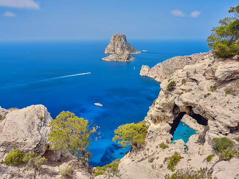Wide-angle view of the iconic islets of Es Vedrà and Es Vedranell, off the southern coast of Ibiza, and an impressive natural arch of rock located on the western cliffs of Cap Llentrisca. Blue sky, picturesque clouds, the dazzling bright light of a summer noon, steep cliffs dotted with Mediterranean scrub, sailing boats, yachts and motorboats crossing crystal-clear waters. High level of detail, natural rendition, realistic feel. Developed from RAW.