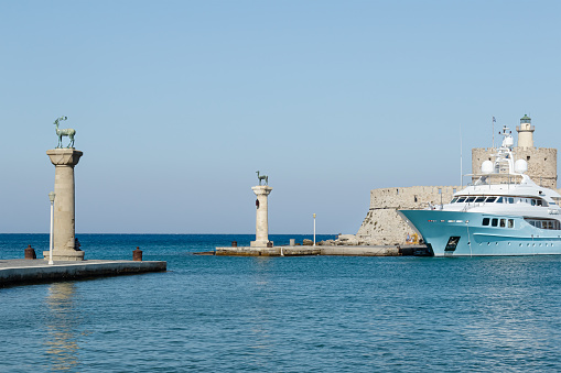 selective focus on columns of the Colossus of Rhodes and Fortress St. Nicholas with  tourist boat in front of it, old harbor entrance