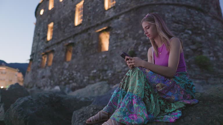 Teenager girl sitting on boulders of Rapallo harbor and using smartphone