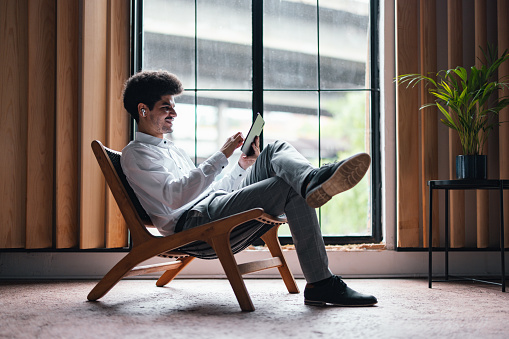 Handsome smiling man sitting in a laid back chair next to a window holding a digital tablet and wearing Bluetooth earbuds. He is having a video call or he is watching something funny on the internet.