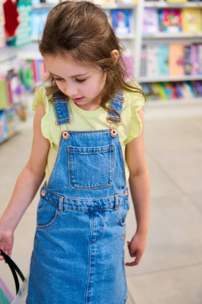 little kid, preschool girl dressed in yellow t-shirt and denim dress, with shopping cart, in the school stationery store - paying children only retail childhood imagens e fotografias de stock