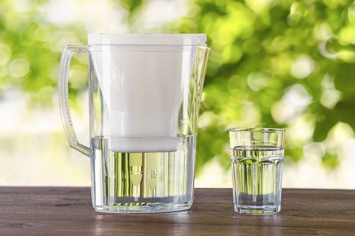Full glass of water with filter jug on wooden table against the background of defocused green leaves