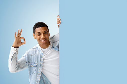 Young African American man peeking out from empty board and shows the OK sign standing on a blue background. Discount, black friday or sale concept