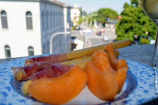 Melon slice with prosciutto crudo in plate at window with Italian urban view