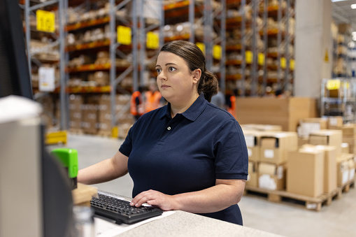 Mid adult woman updating stock on computer in a large logistics facility. Woman working on computer in distribution warehouse.