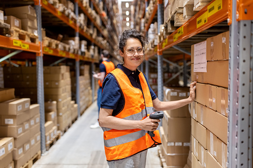 Smiling woman wearing reflective clothing working at distribution warehouse. Happy female warehouse worker looking at camera while scanning boxes on storage rack.