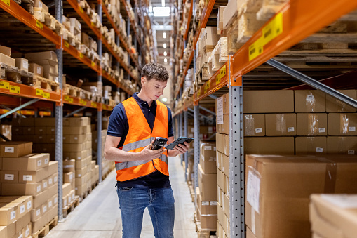 Warehouse worker holding digital tablet and scanner checking cargo on shelves. Male worker in uniform scanning boxes on shelves at distribution warehouse.