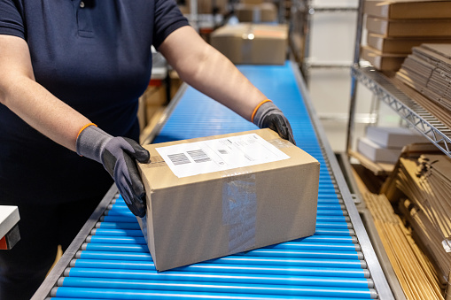 Cropped shot of a female worker wearing gloves placing a parcel on conveyor belt at warehouse.