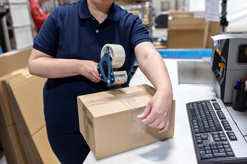 Close-up of a female worker packing delivery package for shipping at logistics center.  Woman working in a warehouse sealing a cardboard box using tape dispenser.