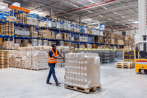 Worker moving boxes on a pallet jack inside a large distribution center. Young African man pushing a pallet jack loaded with boxes in a big storage area.