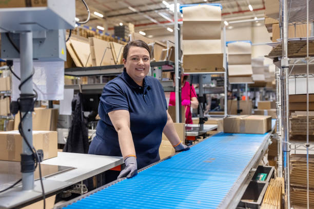 Smiling young woman standing by conveyor belt in distribution warehouse. Female worker working on conveyor belt in packaging plant.
