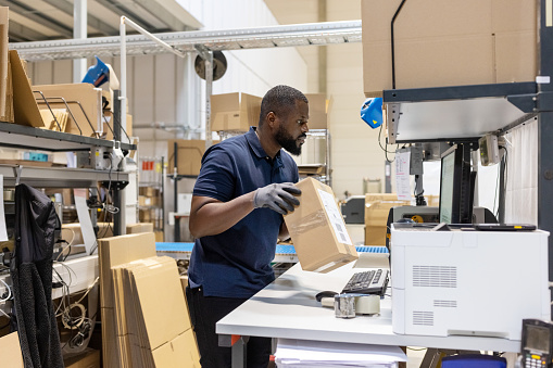 Side view of african man holding a parcel and looking at computer screen working in a distribution warehouse, Young man working in logistics center preparing parcel for delivery.