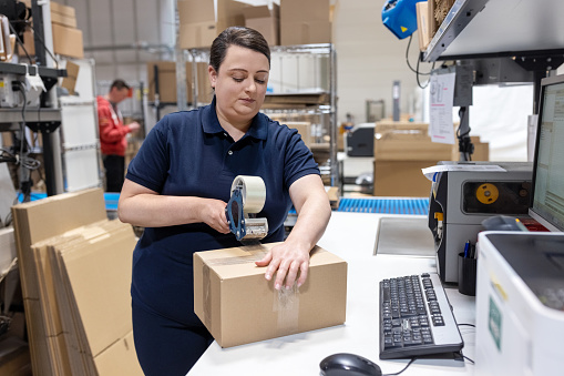 Woman working in distribution warehouse preparing a package for delivery. Female worker packing the goods for shipping to the customer in a cardboard box. Sealing a box with tape dispenser.