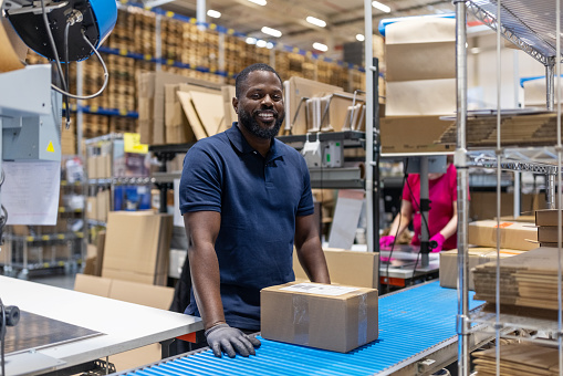 Portrait of smiling young man working in large distribution warehouse standing by conveyor belt. African man working at logistics center looking at camera and smiling.