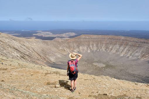 Back view of unrecognizable woman in hat with backpack standing on top of ridge of dry mountains with crater in sunlight