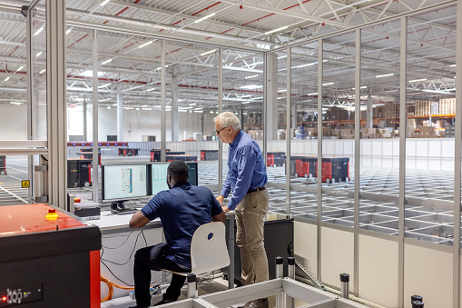 Rear view of man operating AGV in control room in warehouse with manager standing by. Manager with a male worker working on computer in AGV control room at warehouse.