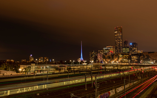 In June 2017, nighttime trains carry passengers to and from Federation Square in the heart of Melbourne, Australia.  In the distance is the pale blue spire sitting atop the Melbourne Arts Center.