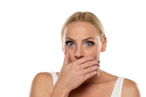 retrato de una mujer rubia avergonzada de mediana edad con maquillaje cubriendo su boca con la mano sobre fondo blanco - women middle aging process covering fotografías e imágenes de stock