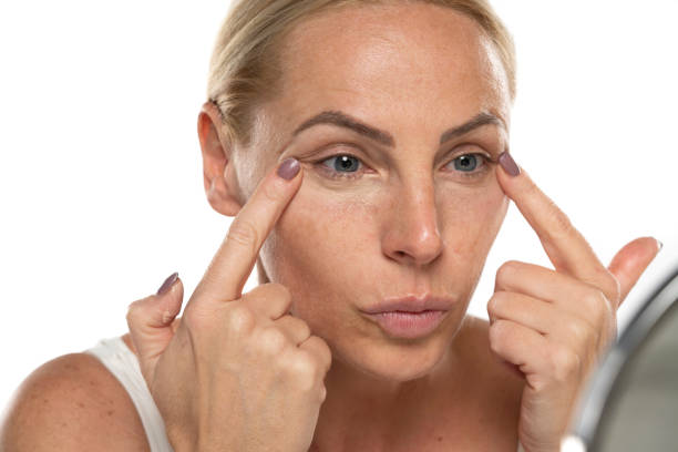 Middle aged senior woman applyes concealer under her eyes on white background